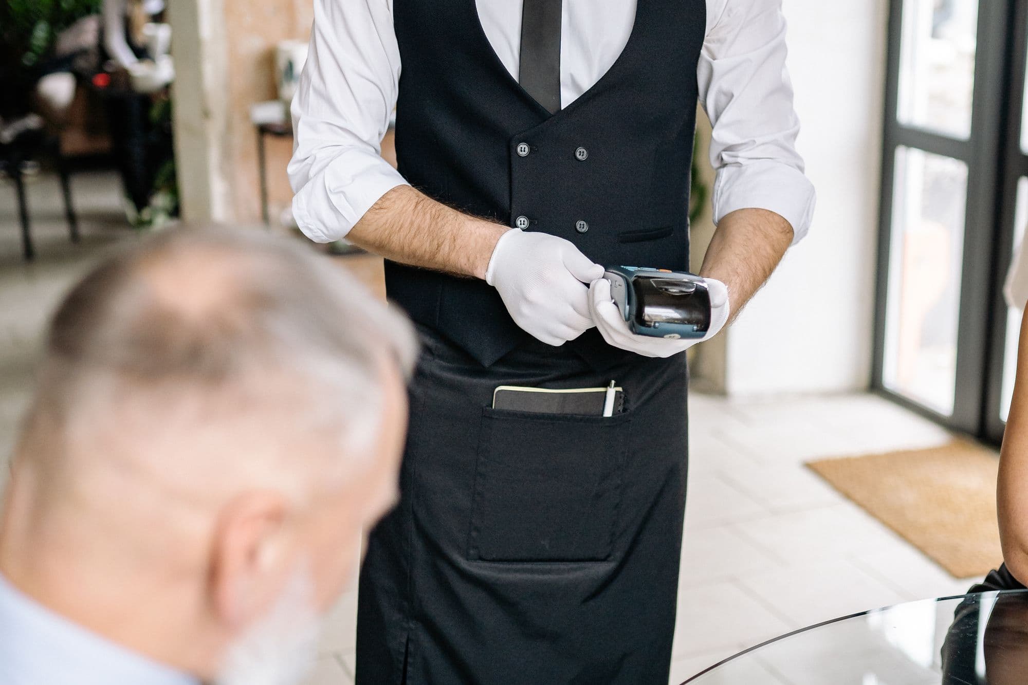 Serveur en gilet noir tenant un terminal de paiement face à un client attablé